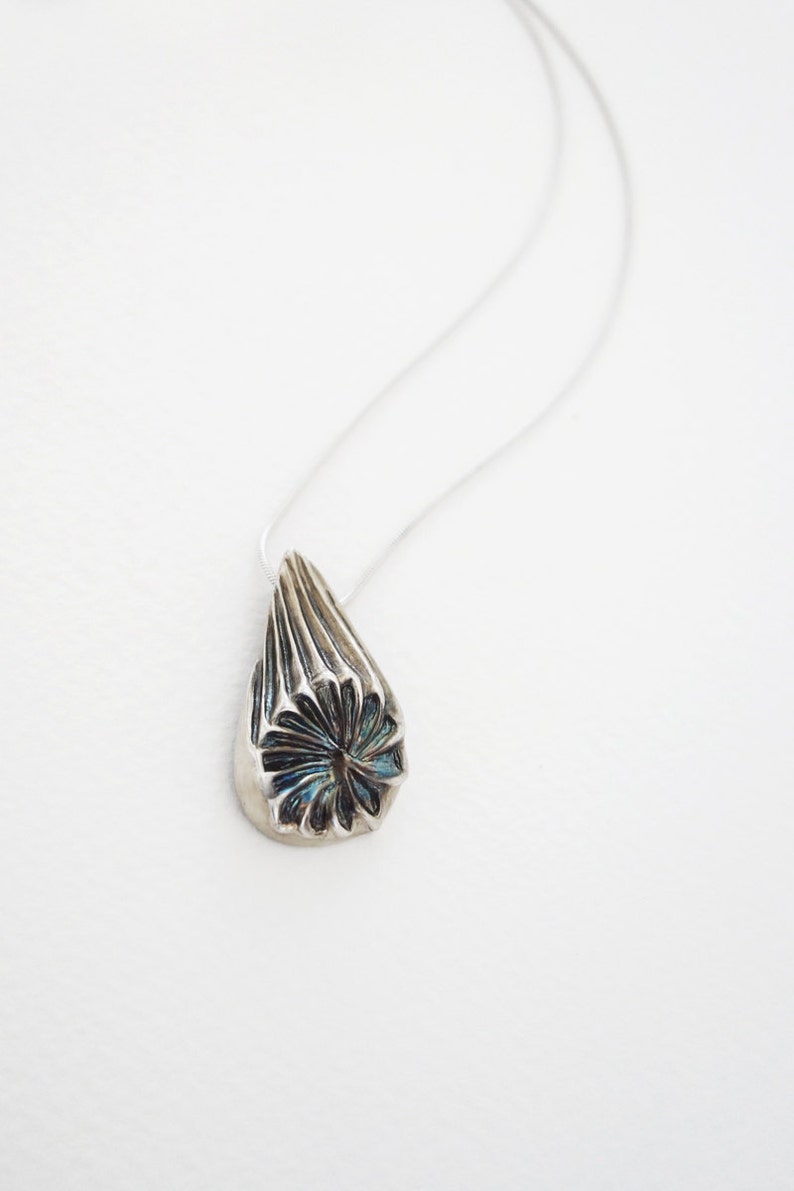 Puede incluir: Un collar de plata con un colgante en forma de l&aacute;grima. El colgante tiene un dise&ntilde;o detallado y texturizado con un centro azul.