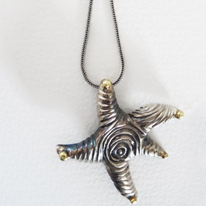 May include: A silver starfish pendant necklace with a textured design and gold accents. The pendant hangs from a silver chain. The starfish has a spiral design in the center and gold tips.