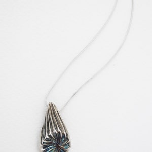 Puede incluir: Un collar de plata con un colgante en forma de l&aacute;grima. El colgante tiene un dise&ntilde;o geom&eacute;trico texturizado con un acabado iridiscente azul y verde.