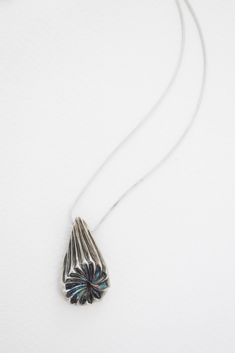 Puede incluir: Un collar de plata con un colgante en forma de l&aacute;grima. El colgante tiene un dise&ntilde;o geom&eacute;trico texturizado con un acabado iridiscente azul y verde.