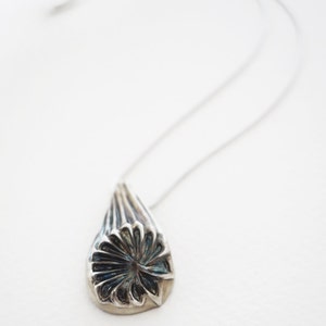 Puede incluir: Un collar de plata con un colgante en forma de l&aacute;grima. El colgante tiene un dise&ntilde;o detallado de una flor con un centro azul.