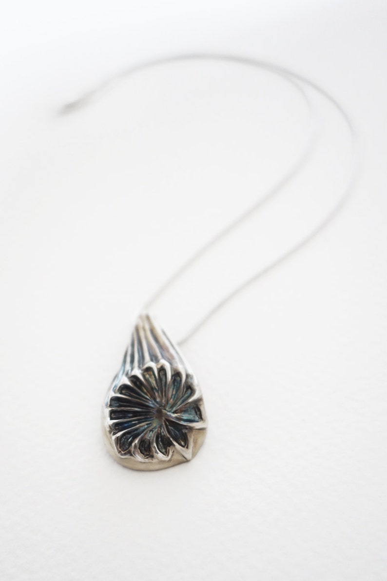 Puede incluir: Un collar de plata con un colgante en forma de l&aacute;grima. El colgante tiene un dise&ntilde;o detallado de una flor con un centro azul.