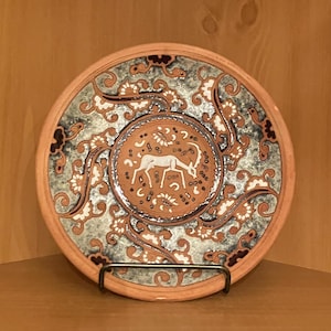May include: A ceramic plate with a silver and white design featuring a deer in the centre. The plate has a brown rim and a blue and white floral pattern.