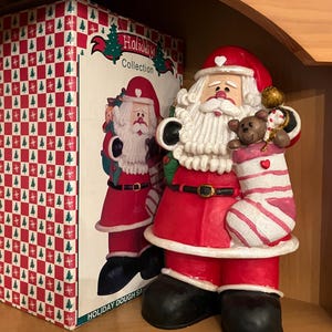 May include: A ceramic Santa Claus figurine in a red suit, black boots, and a white beard, holding a stocking with a teddy bear. The figurine is next to a box with a matching Santa Claus image and the words "Holiday Collection".
