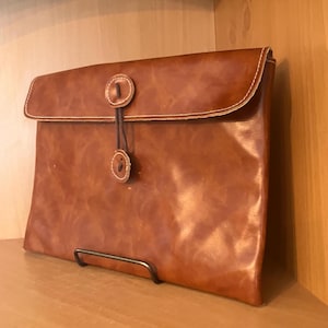 Puede incluir: Un portafolio de cuero marrón con cierre de solapa y detalle de botón. El portafolio rectangular presenta costuras de color claro alrededor de los bordes. El cierre consta de dos botones conectados por un cordón de cuero.