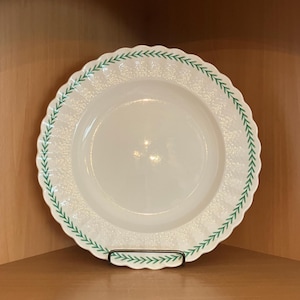 May include: A white ceramic plate with a scalloped edge and a green leaf pattern around the rim. The plate is displayed on a black metal stand. The plate has a textured surface.