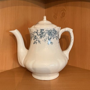 May include: A white ceramic teapot with a blue floral design. The teapot has a lid and a handle. It is sitting on a wooden surface.