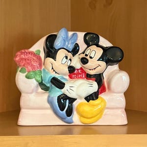 May include: A ceramic figurine featuring Mickey and Minnie Mouse embracing on a pink couch. Minnie wears a blue dress and bow, while Mickey wears red shorts. A bouquet of pink flowers and a heart-shaped sign that says "BE MINE" are also included.