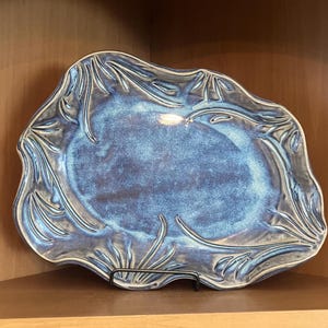 Puede incluir: Un plato de cerámica azul hecho a mano con una forma orgánica única. El plato presenta un acabado brillante y está adornado con un patrón en relieve en forma de hojas alrededor de los bordes. Descansa sobre un soporte de metal negro.