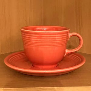 May include: A coral-colored teacup and saucer set. The cup has a rounded shape with a handle and horizontal ridged detailing. The saucer is round with a matching ridged design. The set is made of ceramic.