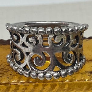 May include: A silver ring with a wide band featuring a delicate, intricate design of swirls and scrolls. The band is adorned with a row of small beads.
