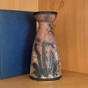 May include: A brown and black ceramic vase with a flared base and a narrow neck. The vase features abstract black brushstrokes and a dark rim. A blue book is partially visible in the background.