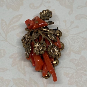Puede incluir: Un broche vintage con un racimo de ramas de coral y flores doradas. El coral es de un color naranja vibrante y las flores tienen un diseño detallado.