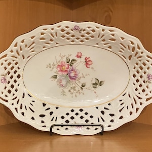 May include: White oval serving dish with a delicate lace design and floral pattern. The dish has a gold rim and is perfect for serving fruit, snacks, or desserts.