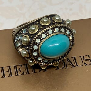 Könnte beinhalten: Ein auffälliger Ring mit einem großen, ovalen türkisfarbenen Stein in der Mitte. Der Ring ist von kleinen, runden, perlenartigen Akzenten und klaren Kristallen umgeben, die in einem goldfarbenen Metall gefasst sind. Der Ring ist von der Marke HEIDI DAUS.