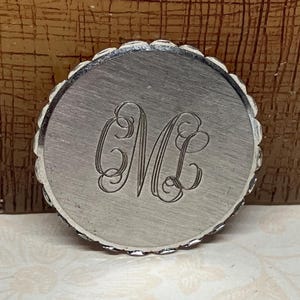 May include: A silver-toned, round object with a scalloped edge. The center features an engraved monogram of the letters "M" and "L" in an elegant, cursive font. The object appears to be a decorative piece or a component of a larger item.
