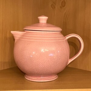 Könnte beinhalten: Eine rosa Keramik-Teekanne mit Deckel und Henkel. Die Teekanne hat einen runden Körper und eine Craquelé-Glasur. Die Teekanne steht auf einem Holzregal.