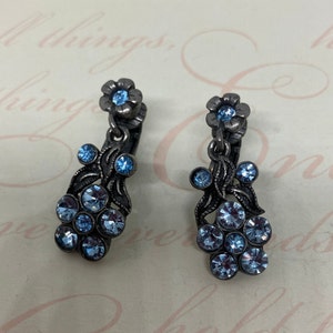 May include: A pair of silver tone clip-on earrings with a floral design. Each earring features a cluster of light blue rhinestones and a single light blue rhinestone flower at the top.