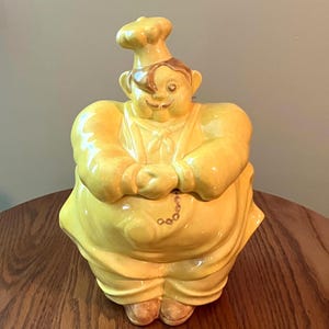 May include: A yellow ceramic cookie jar shaped like a chef wearing a tall toque, a neckerchief, and an apron. The figure has brown hair, a mustache, and hands clasped in front. A small brown chain detail is visible on the front of the apron.