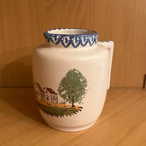 Pode incluir: Jarro de cerâmica branca com uma borda azul e branca ao redor da parte superior. O jarro tem uma alça e apresenta uma cena pintada à mão de uma aldeia com uma árvore verde.