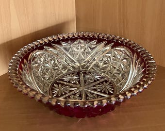 May include: A clear glass bowl with a ruby red base and rim. The bowl features a detailed starburst and geometric pattern. The rim has a scalloped edge. This decorative bowl is ideal for serving or display.