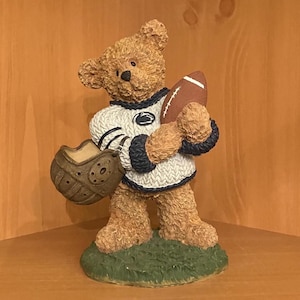 May include: A brown teddy bear figurine wearing a white and navy blue sweater and holding a brown football. The bear also holds a brown football glove. The figurine is standing on a green base.