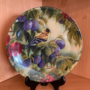 May include: A round glass plate with a bird and fruit design. The plate features a yellow bird with black markings perched on a branch with purple plums and red berries. The plate is on a black stand.
