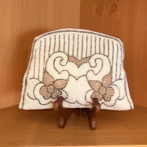 May include: A white beaded clutch purse featuring vertical silver stripes and a symmetrical floral pattern in gold and white beads. The bag has a curved top edge and a zipper closure along the side, showcasing intricate beadwork throughout the exterior.