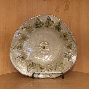Op de afbeelding: Een beige keramische schaal met een bloemenmiddelpunt en een rand van groene en beige klimopbladeren. De schaal wordt gepresenteerd op een zwarte metalen standaard, die de handgemaakte kwaliteit en rustieke charme benadrukt.