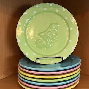Puede incluir: Una pila de platos de cerámica coloridos con un plato verde encima. El plato verde tiene un diseño de pájaro y lunares blancos. Los platos se exhiben en una estantería de madera.