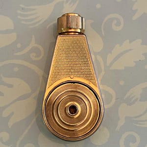 Könnte beinhalten: Ein goldfarbenes Feuerzeug aus Metall mit tropfenförmigem Gehäuse, das im oberen Bereich ein texturiertes Kreuzschraffurmuster und im unteren kreisförmigen Bereich eine Reihe konzentrischer Kreise aufweist. Oben befindet sich ein kleiner Knopf.