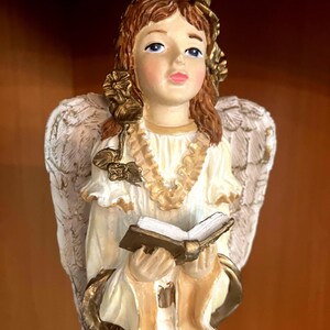 May include: A ceramic angel figurine with a serene expression, wearing a white and gold gown, and holding an open book. The angel has feathered wings and a halo. The figurine is painted with soft colors and gold accents.