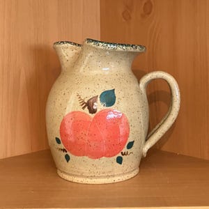 May include: A ceramic pitcher with a speckled cream-colored glaze, featuring a hand-painted orange pumpkin design on the front. The pitcher has a curved handle, a wide pouring spout, and a dark green speckled rim detail on the top edge.