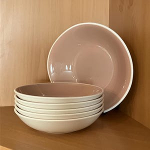 Pode incluir: Um conjunto de seis tigelas de cerâmica branco-sujo e malva, com uma borda larga, empilhadas. Uma tigela maior está atrás da pilha. As tigelas estão em uma prateleira de madeira, adequadas para uma cozinha ou sala de jantar.