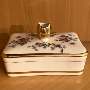 Puede incluir: Una caja de porcelana blanca con ribete dorado y diseño floral. La tapa tiene un pájaro dorado y un pomo en forma de tulipán dorado.