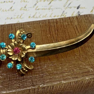 May include: A vintage gold-toned brooch in the shape of a curved sword. The brooch features a floral design with a pink center stone and teal blue accent stones. The brooch is set on a brown surface.
