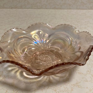 May include: A pink iridescent glass bowl with a scalloped edge and a floral design. The bowl is made of clear glass with a pink iridescent finish.