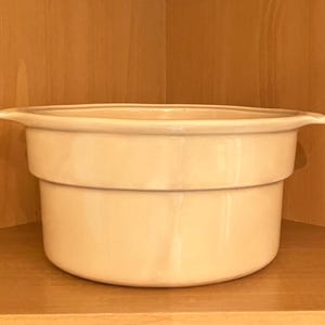 May include: A light beige ceramic casserole dish with a rounded base and a wide, flat rim. The dish has a two-tiered design and is set against a wooden background. The overall shape is circular, and the dish appears to be empty.