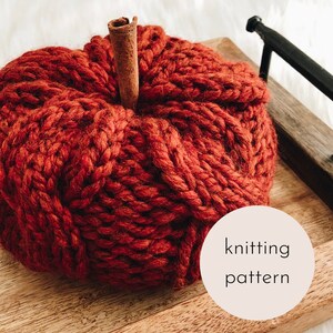 Könnte beinhalten: Ein gestrickter Kürbis in einem tiefen Rot mit einem Zimtstangen-Stiel. Der Kürbis liegt auf einem Holztablett mit dem Text "knitting pattern" auf einem weißen Kreis.