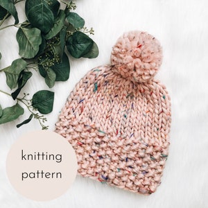 May include: A pink knitted hat with a pom pom on top. The hat has a speckled pattern with multi-colored flecks throughout. The hat is on a white surface with a green leafy branch in the background. The text "knitting pattern" is written on a white circle.