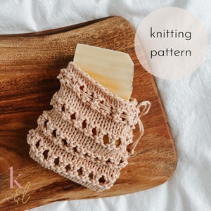 Op de afbeelding: Een gebreide zeepzak met een stuk zeep erin. De zak is lichtroze en heeft een gehaakt ontwerp. De zeepzak ligt op een houten snijplank. De tekst "knitting pattern" staat op een witte cirkel in de rechterbovenhoek van de afbeelding.