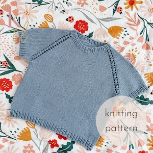 May include: A light blue knitted jumper with a round neckline and short sleeves. The jumper is laid out on a floral fabric background. The text "knitting pattern" is written in white on a circle in the lower right corner of the image.