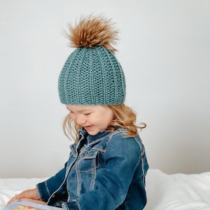 May include: A young child wearing a teal blue knitted beanie with a brown faux fur pom pom sits on a white bed and reads a book. The child is wearing a blue denim jacket.