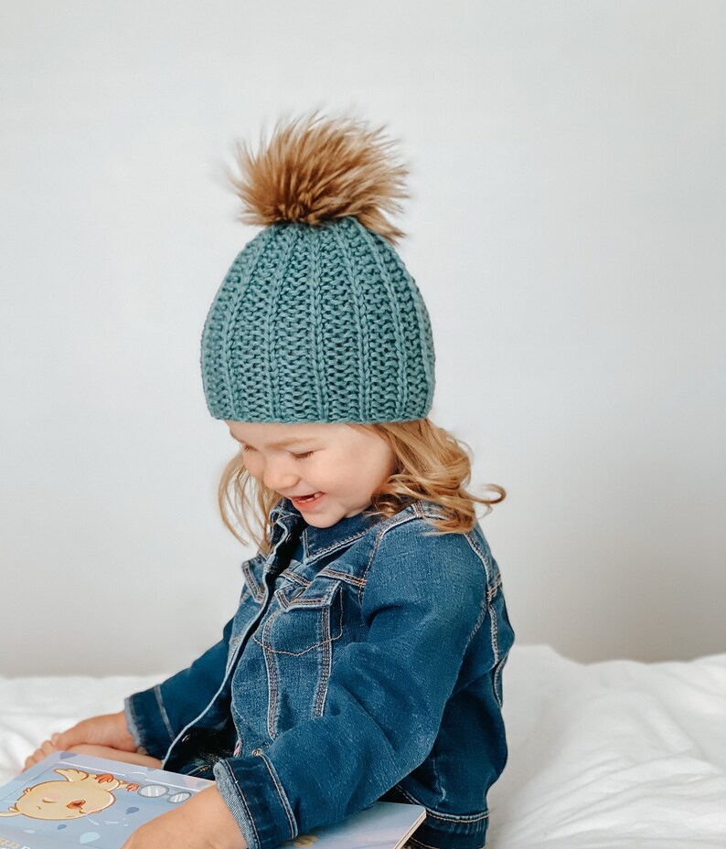 May include: A young child wearing a teal blue knitted beanie with a brown faux fur pom pom sits on a white bed and reads a book. The child is wearing a blue denim jacket.