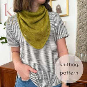 May include: A knitted olive green triangular shawl is worn over a striped t-shirt. The shawl has a textured appearance with a decorative edge. The image includes the text "knitting pattern" in a circular graphic.
