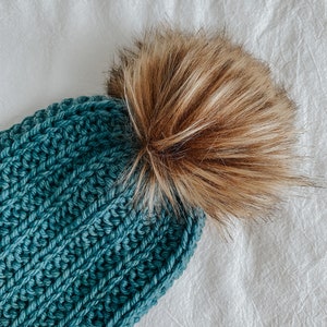 May include: A teal blue knitted beanie with a brown faux fur pom pom.