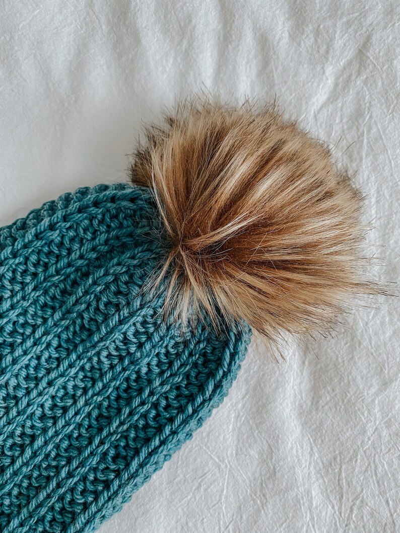 May include: A teal blue knitted beanie with a brown faux fur pom pom.