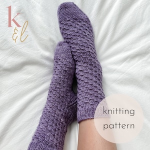 Op de afbeelding: Een paar paarse gebreide sokken met een textuurpatroon. De sokken liggen op een wit beddengoed. De tekst "knitting pattern" is zichtbaar in de rechterbenedenhoek van de afbeelding.