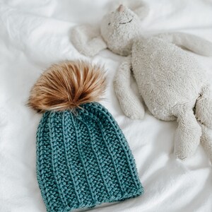 May include: A teal blue knitted beanie with a brown faux fur pom pom. The beanie is lying on a white bedspread next to a white stuffed animal.