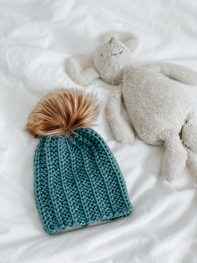 May include: A teal blue knitted beanie with a brown faux fur pom pom. The beanie is lying on a white bedspread next to a white stuffed animal.